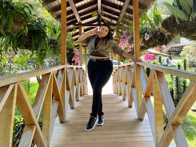 MissSofiia with tits covered, wearing a green tie-front top, black jeans, and black sneakers, stands on a wooden bridge, hand in hair, under a wooden roof.