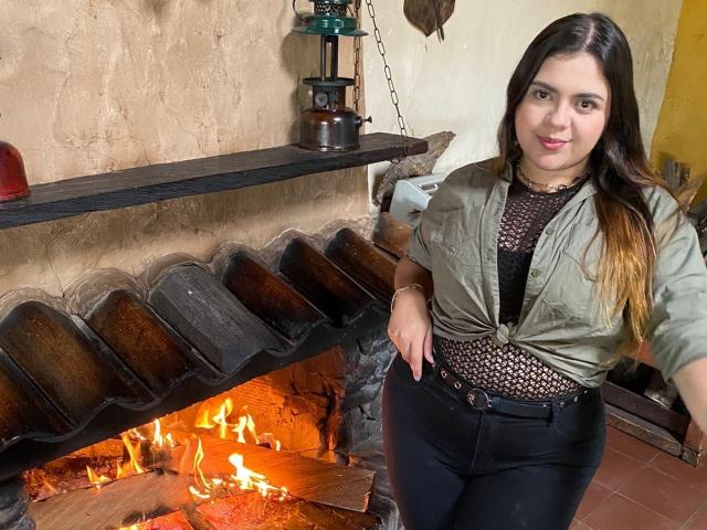 MissSofiia with long brown hair, green jacket, black patterned shirt, and black pants, standing by a burning fireplace, smiling at the camera, showing a cute ass.