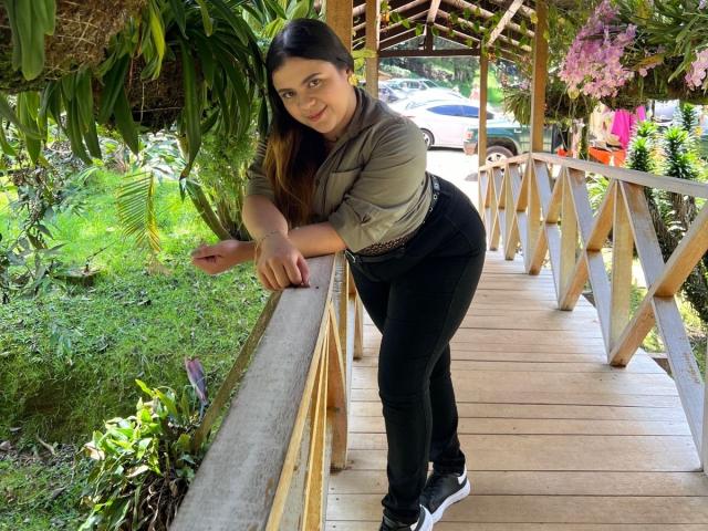 MissSofiia with long dark hair, green shirt, black jeans, and black shoes, leaning on a wooden bridge, smiling, her ass slightly raised, surrounded by greenery and pink flowers.
