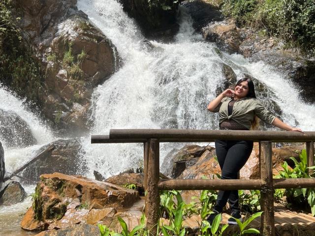 MissSofiia with her tits and ass standing by a wooden railing, wearing a green shirt and black pants, in front of a flowing waterfall. She's touching her face.