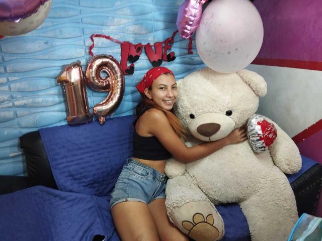CamilaaEvans, with long brown hair and red bandana, hugs a huge white teddy bear, wearing a black crop top and blue shorts, sitting on a blue couch.