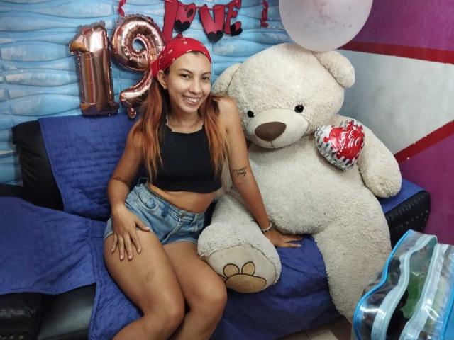 CamilaaEvans with red bandana, black sports bra, and denim shorts, sitting next to a huge white teddy bear holding a red heart, on a blue-covered couch.