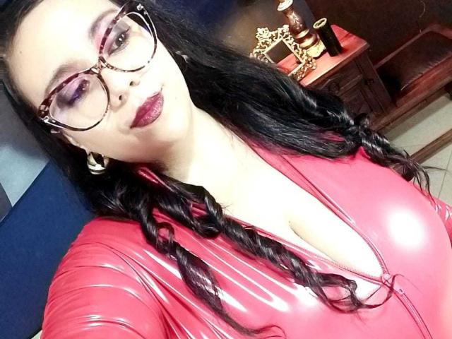 LexyDom with big glasses, red latex dress, and braided black hair, showing off her tits in a selfie. She's got dark lipstick and a sexy, confident look.