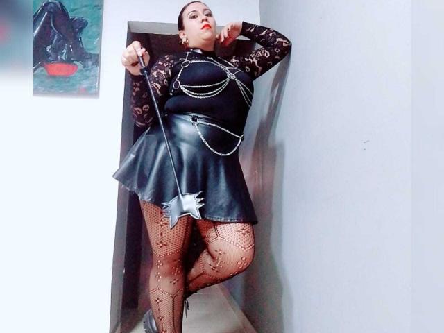 LexyDom, with large tits, wearing a black lace top, leather skirt, and fishnet stockings, holds a whip, standing in a hallway, her red lipstick contrasting her pale skin.