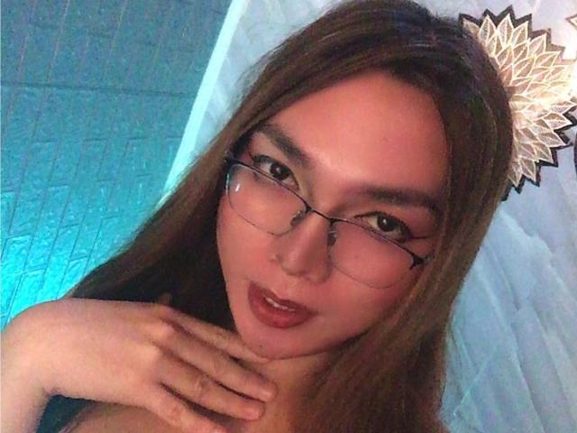 AsiandollBella with glasses, brown hair, and glossy red lips, touching her neck, showing her soft tits and a cute, slightly open-mouthed expression in a selfie.