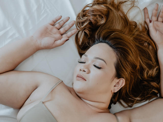 DelightfulCurvy with closed eyes, lying on white bed, arms above head, brown hair spread, wearing beige top, soft smile, fair skin, small earrings, relaxed and peaceful.