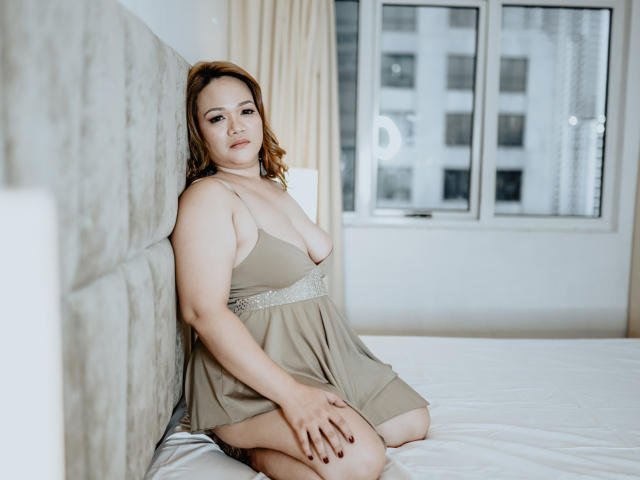 DelightfulCurvy, a plus-sized woman with pale skin and red hair, sits on a bed in a beige nightie, showing her small tits and resting her hand on her thigh.