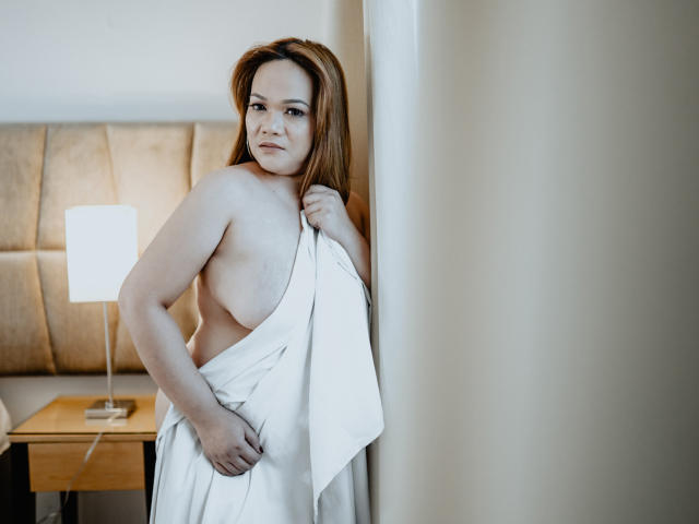 DelightfulCurvy with pale skin, red hair, and small tits, stands in a room, partially covering her naked body with a white towel, holding it against her pussy.