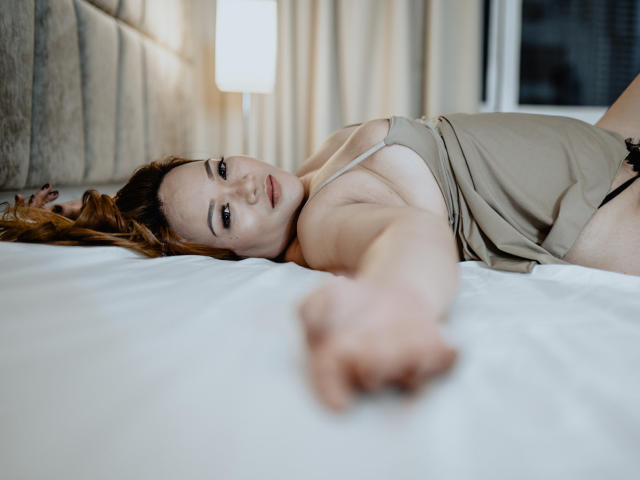 DelightfulCurvy with tits visible, lying on a bed, reaching out, wearing a beige top, brown hair, looking at the camera, white sheets, and a blurred lamp in the background.