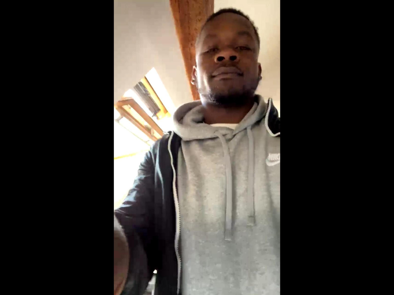 MthHot, a black dude with a short beard, wearing a gray hoodie and black jacket, smiling slightly, standing in a dimly lit room, shot from a low angle.