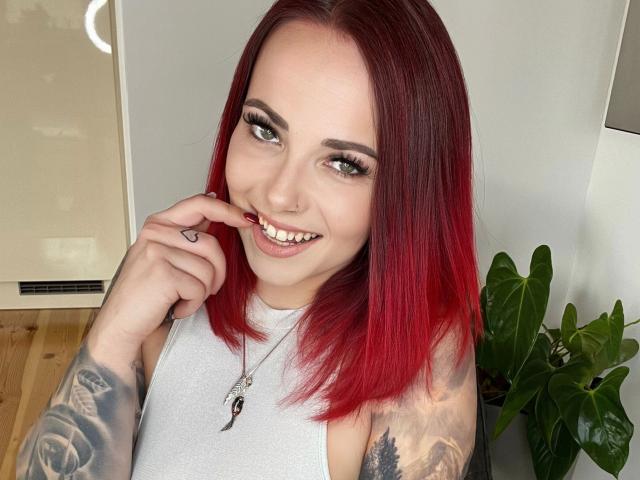 NinaDevil - cheveux roux, souriante, montrant ses dents, touchant son nez avec son doigt, haut blanc sans manches, tatouages sur les bras, yeux verts, anneau au nez, collier avec un pendentif "9", sol en bois..