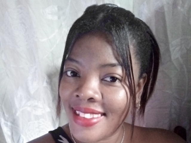 BrownieBabe has dark skin, black hair in a ponytail, red lipstick, and a smile showing white teeth. She wears a black and white top, small hoop earrings.