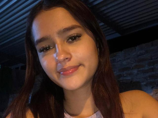 Melody Mansson tem cabelo castanho, olhos verdes e um sorriso adorável com lábios rosados, vestindo uma blusa sem mangas, em um quarto com pouca luz, com tons de azul. Suas nádegas não estão visíveis..