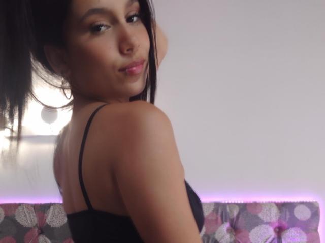 KataleyaMansson with dark hair, looking over her shoulder, wearing a black strap top, showing her bare back and a slight smile, with purple light in the background.