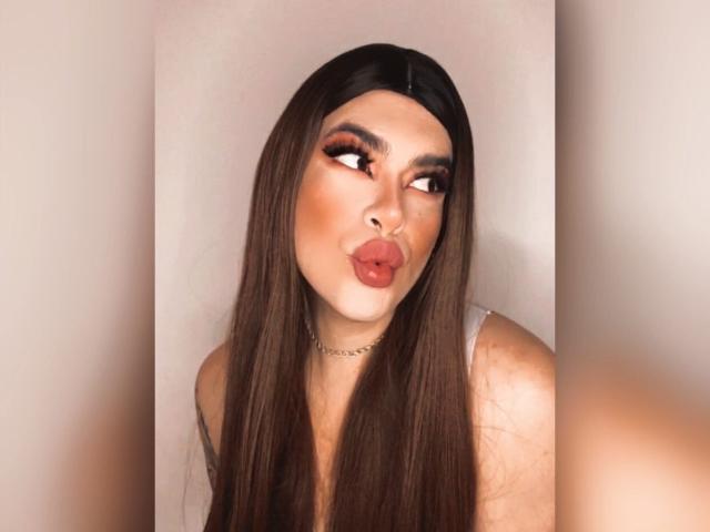 ImNicol with long brown hair, pouty red lips, and dramatic orange eyeshadow, wearing a sheer top and a chain necklace, looking to the side.