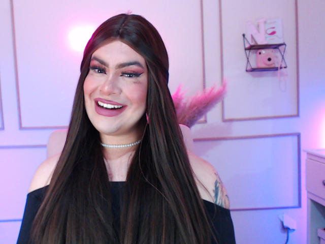 ImNicol met lang bruin haar, lachend, draagt een zwarte top met open schouders, heeft roze oogmake-up, een parelketting, een tatoeage op de rechter schouder, en de belichting is roze-blauw..