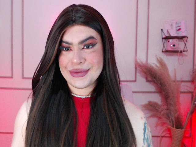 ImNicol has long brown hair, red lipstick, and a cute smile. She's wearing a red top and a silver necklace. Her makeup is bold with pink eyeshadow.