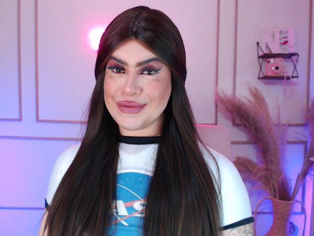 ImNicol has long dark brown hair, full makeup with pink eyeshadow, and pink lipstick. She's wearing a white and blue shirt, smiling slightly. Her tattoo is partially visible.