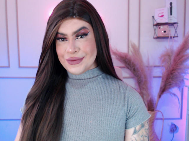ImNicol with long brown hair, gray turtleneck, pink eyeshadow, and tattoos on her arm, smiling at the camera, pink and blue lighting.