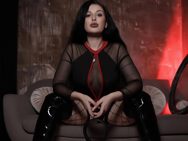 KellyKent-hot with long black hair, wearing sheer black top, red lines, black fishnet, and shiny black boots, sitting on a gray couch with hands on knees, showing her pussy.