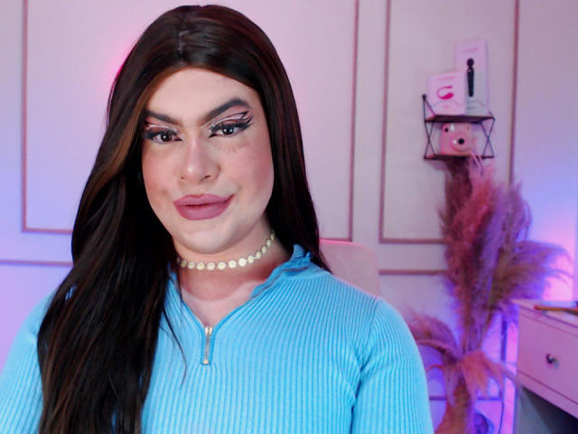 ImNicol with long brown hair, blue ribbed zip-up, pearl choker, and eyeliner, smiling at the camera. Pink and purple lighting on their face. Boobs not visible.