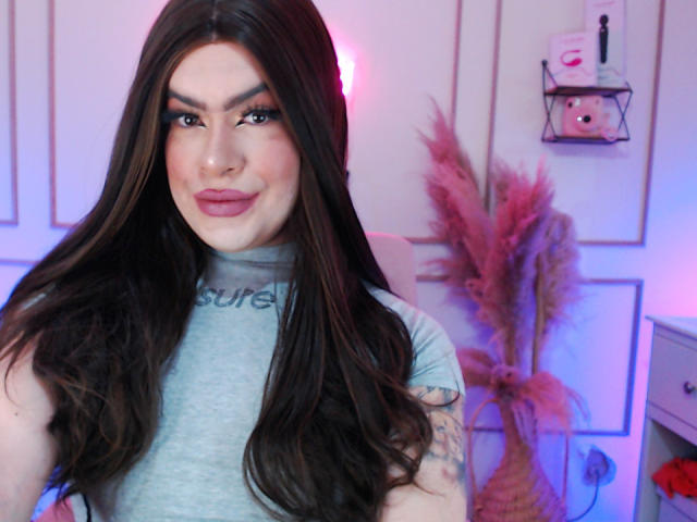 ImNicol with long brown hair, gray t-shirt, and tattoo on right arm, smiling with pink lipstick, blue eyes, and subtle makeup, standing in a room with pink and blue light.