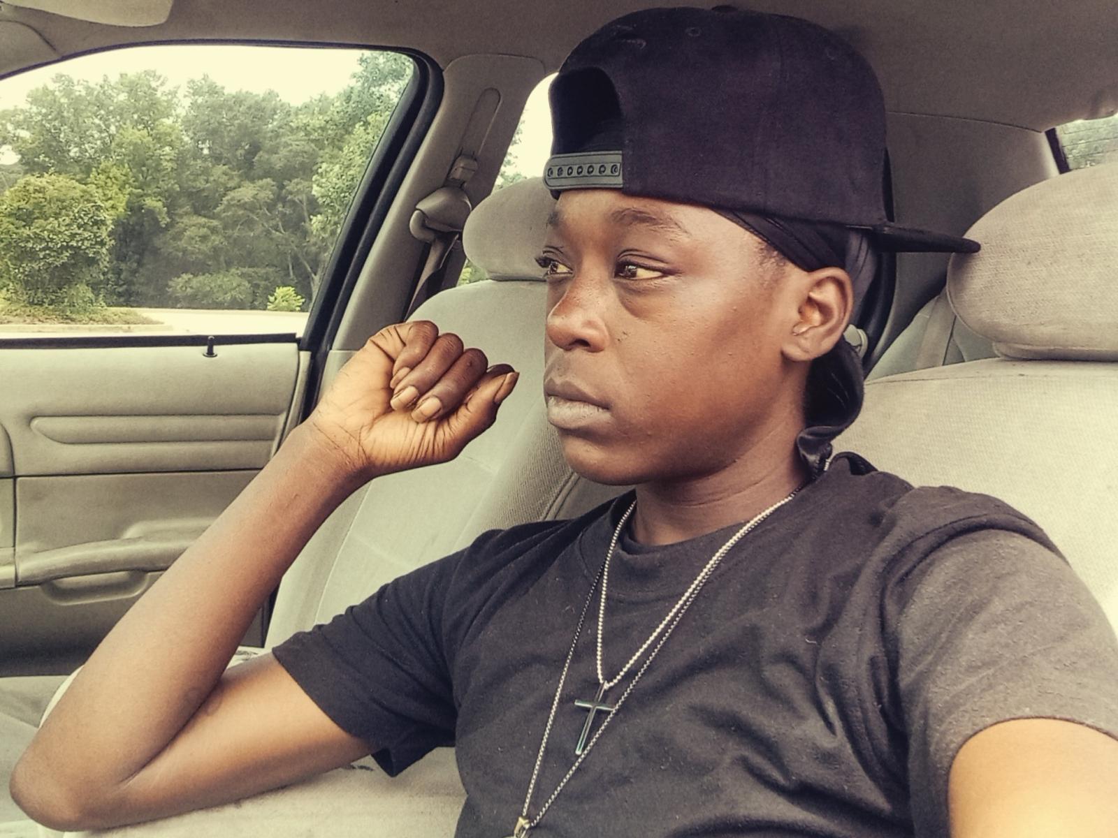 LionessLeo, a dark-skinned dude, wearing a black cap, black t-shirt, and a cross necklace, sits in a car, looking serious with his hand near his face.