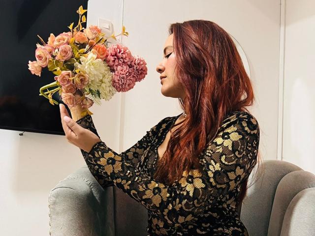 Eine Frau mit roten Haaren, die ein schwarzes Spitzenkleid trägt, einen Strauß aus rosa und weißen Blumen hält und auf einem grauen Stuhl sitzt, während sie die Blumen betrachtet..