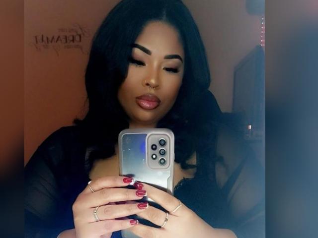 ChineReid69x with dark wavy hair, nose ring, and glossy pink lips, holding a phone with red nails, wearing a black sheer top, taking a selfie.