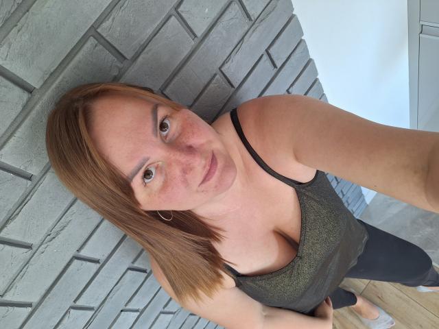 MartyNusiak with brown hair, freckles, green tank top showing big tits, black straps, black pants, standing against gray brick wall, looking up at camera.