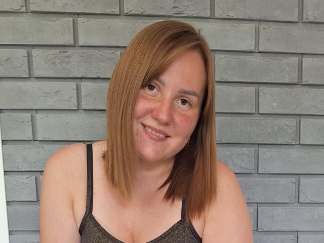 MartyNusiak has shoulder-length brown hair, smiling with freckles, wearing a black spaghetti-strap top, showing her small tits and pale skin.