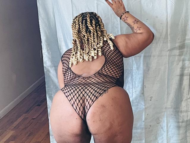 BigMara with blonde braided hair, wearing a black fishnet bodysuit, facing away, showing her big ass and back tattoo of stars. She's standing in a room with wooden floors.