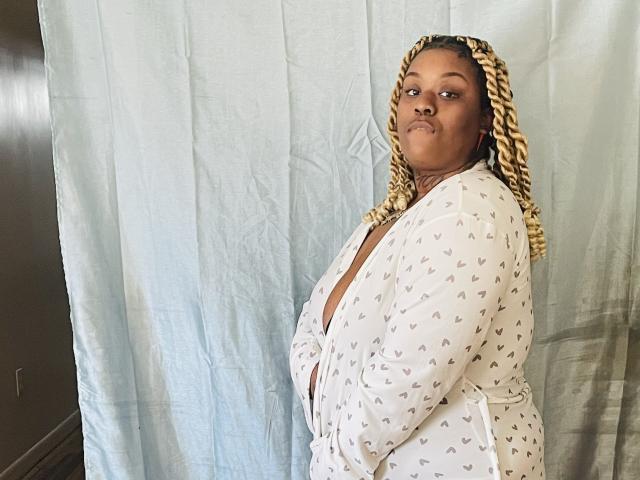 BigMara, a plus-sized black woman with braided blonde and brown hair, wears a white heart-patterned robe, looking slightly to the side with a subtle pout.
