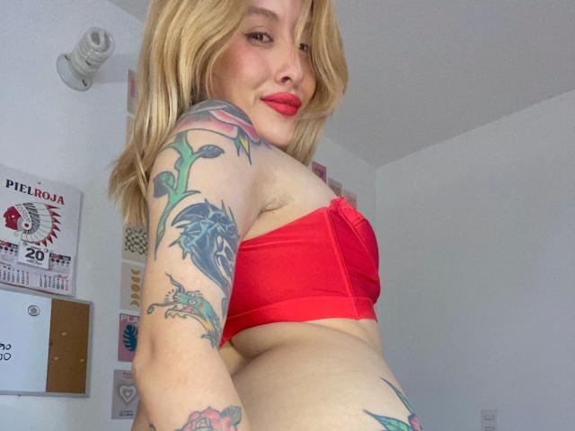 IsabellaPalacio with blonde hair, red bra, and tattoos, showing her tits and ass, smiling with red lipstick, in a white room.