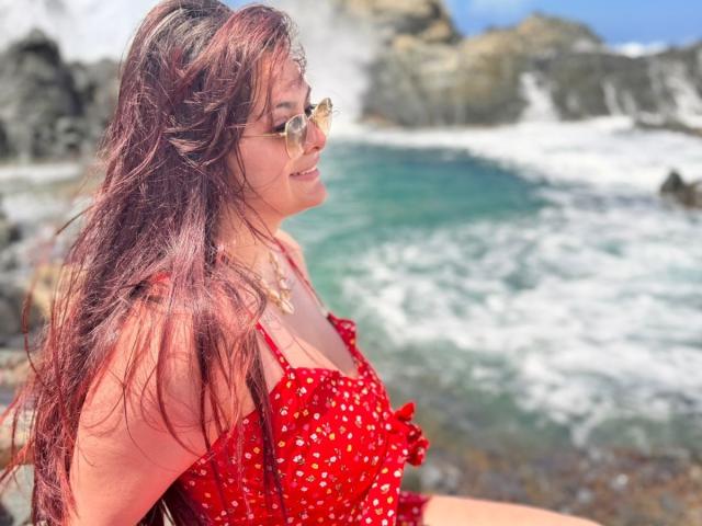 KatherinLatin with red polka-dot dress, glasses, and wet brown hair, showing her tits, standing by a rocky, wave-crashing shoreline.