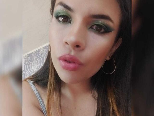 VioletaStone com sombra verde cintilante, batom rosa e brincos de argola, olhando diretamente para a câmera com uma expressão levemente emburrada, cabelo castanho com pontas em degradê e pele clara..