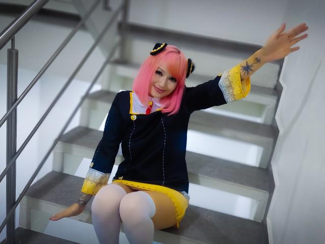 SarahRousen with pink hair, black and yellow cosplay outfit, white thigh-highs, sitting on stairs, raising right arm, tattoo on left forearm, smiling, showing her ass.