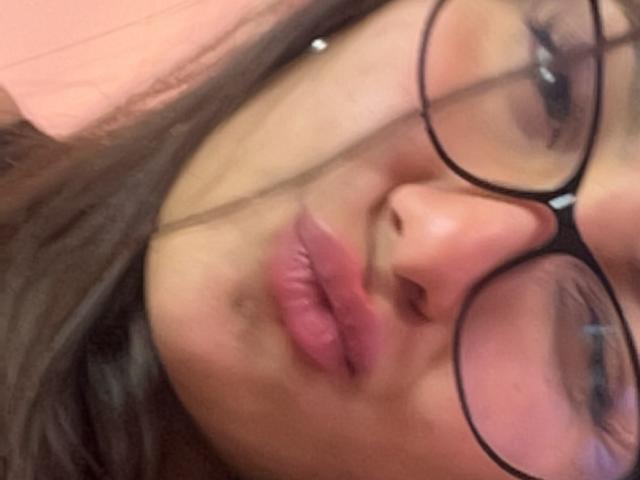 DianaFernandezz's face with puffy pink lips, wearing black glasses, and brown hair, showing a slight pout. Her nose is centered, and she has a small silver nose stud.