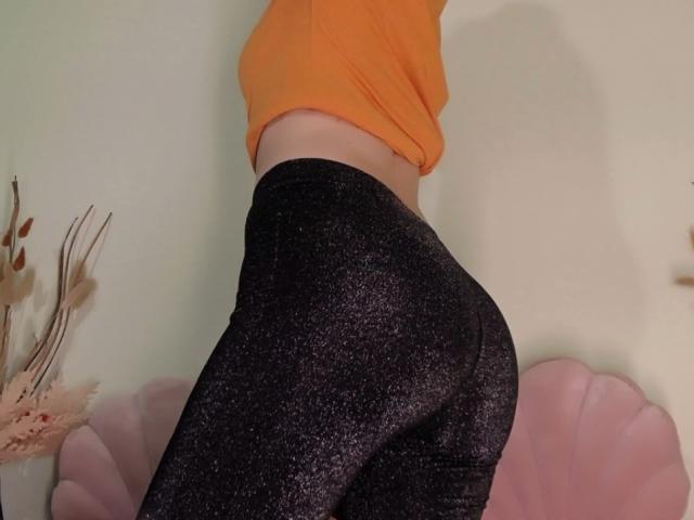 BlueNightX with a small orange top, wearing sparkly black booty shorts, showing their curvy ass and tight butt in a side profile. Pink, fluted chair in the background.