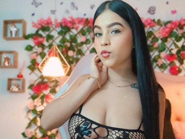 LucyCute with long black hair, pouty lips, and tan skin, wearing a black fishnet top, showing her big tits, touching her neck, in a softly lit room.
