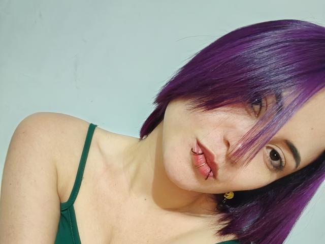 CassieJones with purple hair, pouty lips, and a lip piercing, wearing a green spaghetti strap top, looking at the camera. Her tits are small, and she's cute.