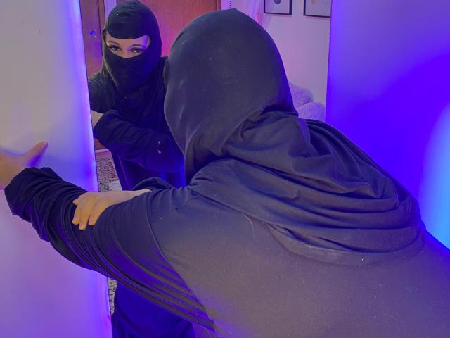 SalmaAzaman in black mask and hoodie, facing mirror, shows finger to reflection, blue lighting, her hands and face visible, her back to camera, her yellow nails.