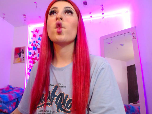 SammyViolet with long red hair, pouting her lips, wearing a gray shirt, has thick eyelashes and is looking up. Her nose is pierced. She's in a pink-lit room.