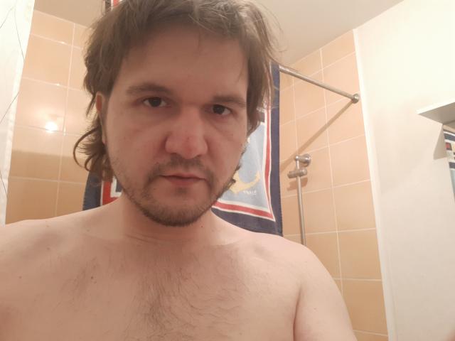 NectarineFad, sem camisa, com um peito levemente gordo e peludo, cabelo castanho ondulado e barba, olhando para a câmera em um banheiro..
