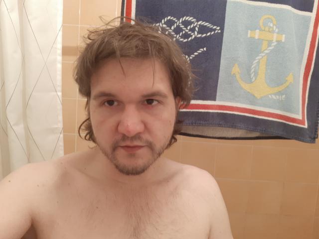 NectarineFad, shirtless, with wet, brown, medium-length hair, and a light beard, has a neutral expression, and is in a bathroom with a blue towel behind him.