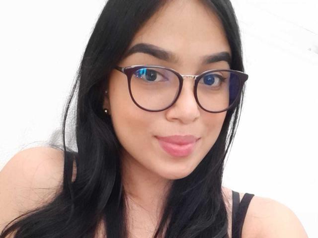 AnyaCathy a les cheveux noirs, des lunettes marron et des lèvres roses légèrement retroussées. Elle porte un haut noir à bretelles qui laisse entrevoir son visage et sa partie supérieure de la poitrine..