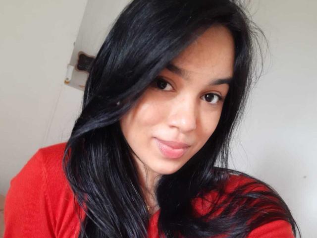 AnyaCathy mit langem, schwarzem Haar, die ein rotes Hemd trägt, hat ein süßes Lächeln und ein leicht gesprenkeltes Gesicht und blickt mit braunen Augen in die Kamera..