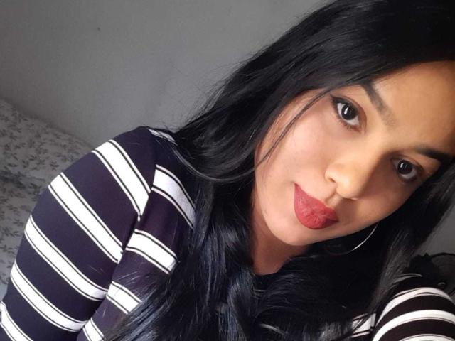 AnyaCathy, avec ses cheveux noirs, son rouge à lèvres rouge, ses boucles d'oreilles et sa chemise rayée noire et blanche, montre son joli visage et ses lèvres pulpeuses. Ses seins ne sont pas visibles..