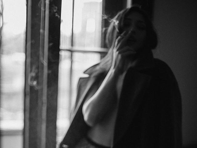MoniFoxy with blurry face, hand near mouth, smoking, topless, wearing open jacket, soft light from window, grayscale, slightly out of focus, sensual, moody.