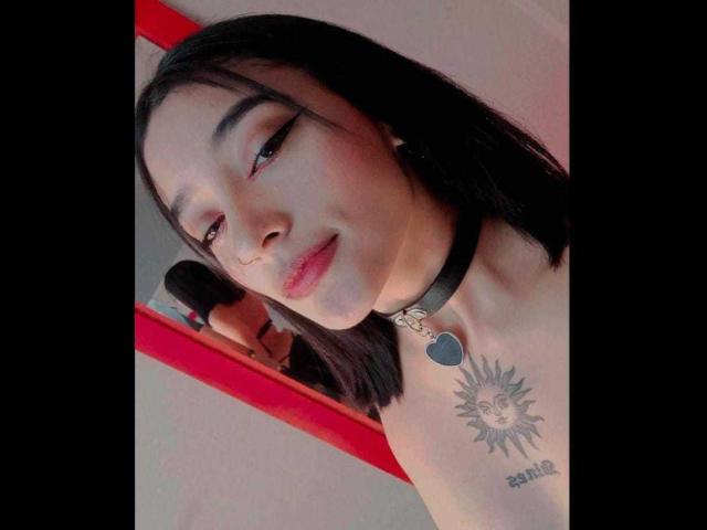 SalomeMille with black hair, pink eyeliner, and red lipstick, wearing a black choker with a heart, showing a sun tattoo on her chest, in a mirror selfie.